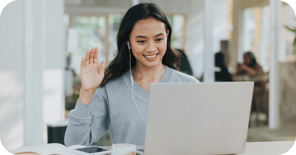 A smiling woman engaged in a virtual meeting, wearing earphones and waving at her laptop screen, representing effective remote team management with strong support systems.